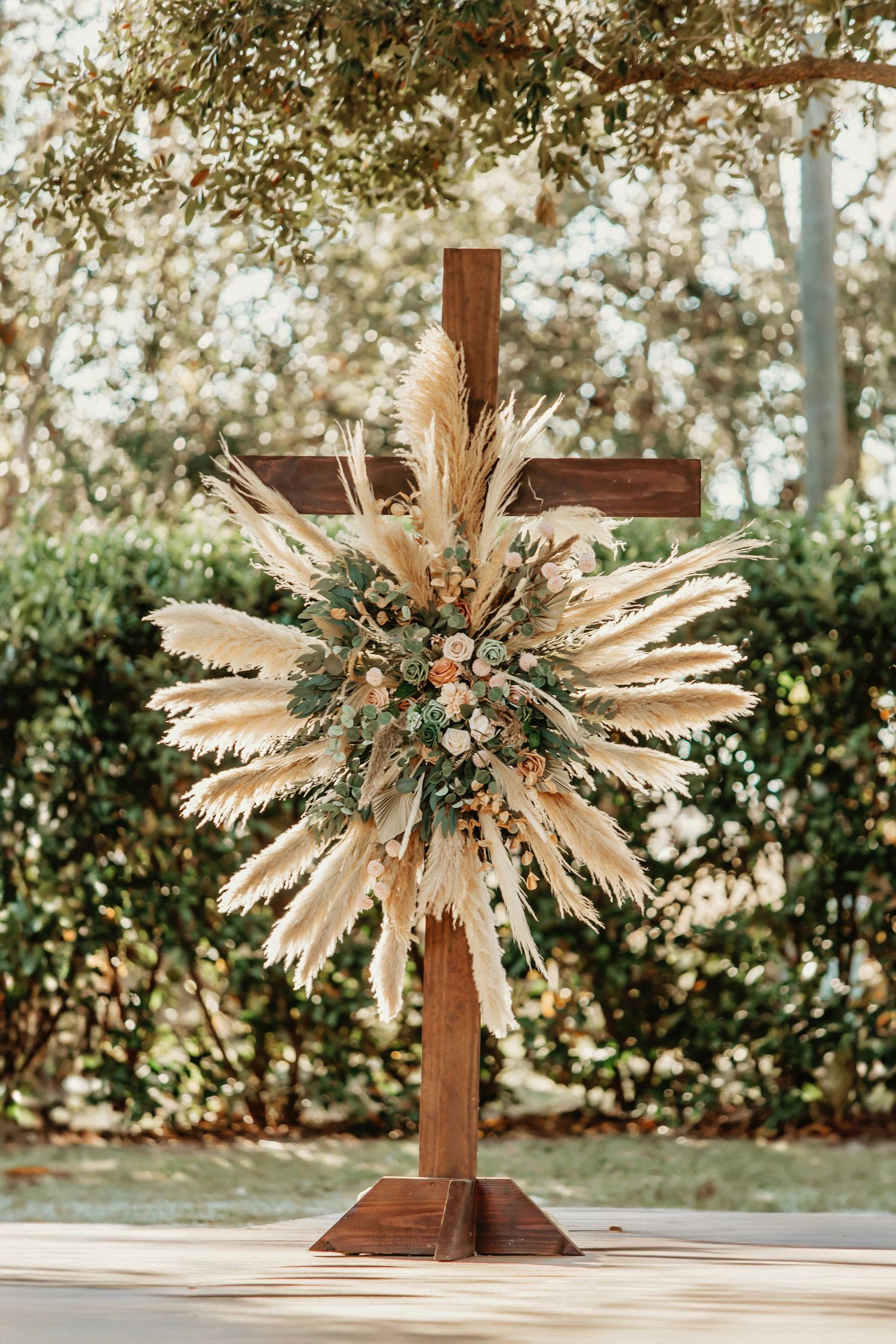 A rustic wooden cross decorated with a large sunburst arrangement of dried pampas grass and neutral florals in an outdoor setting.