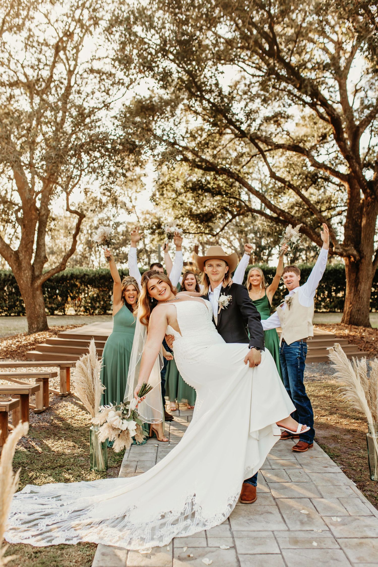 A joyful wedding party celebrates on a paved garden path surrounded by trees.