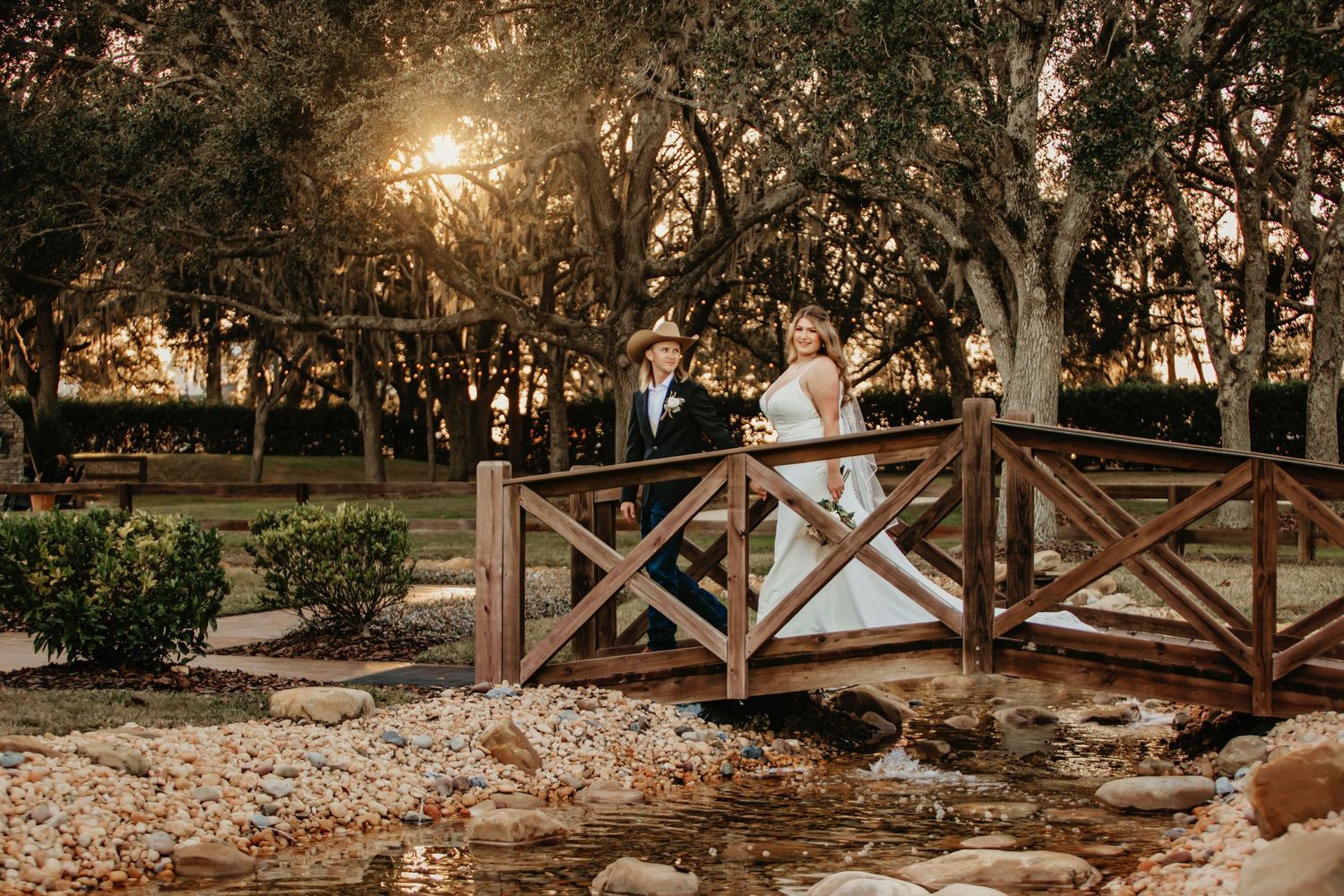 A romantic sunset photograph on a wooden bridge surrounded by lush trees and gardens.