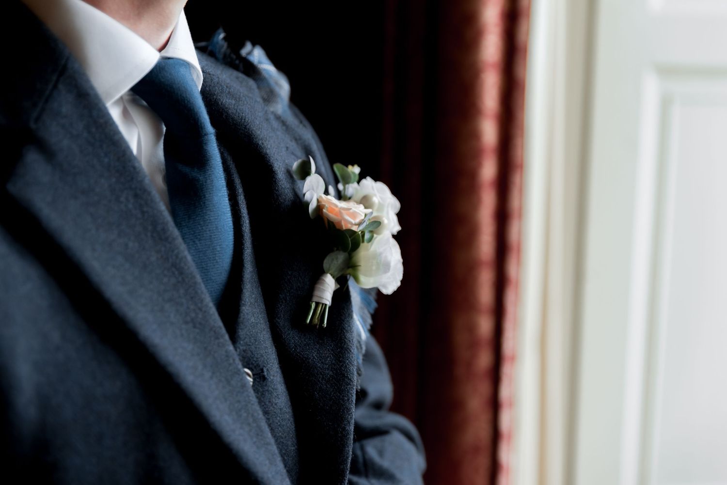 A white rose and greenery boutonniere pinned to a formal navy blue suit lapel for a wedding.