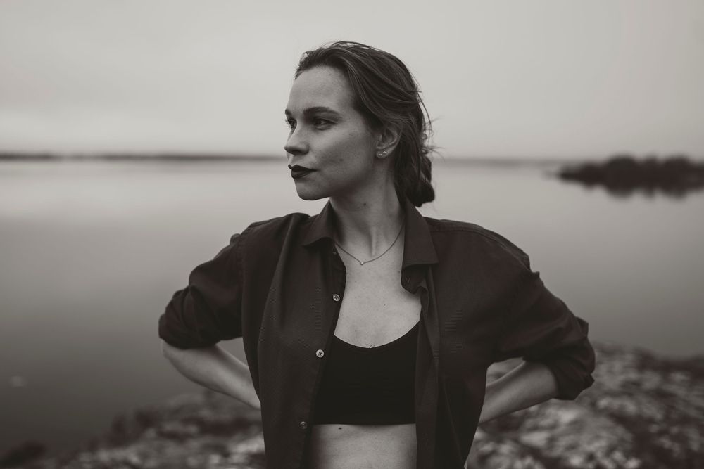 Moody black and white portrait with water and cloudy sky in background.