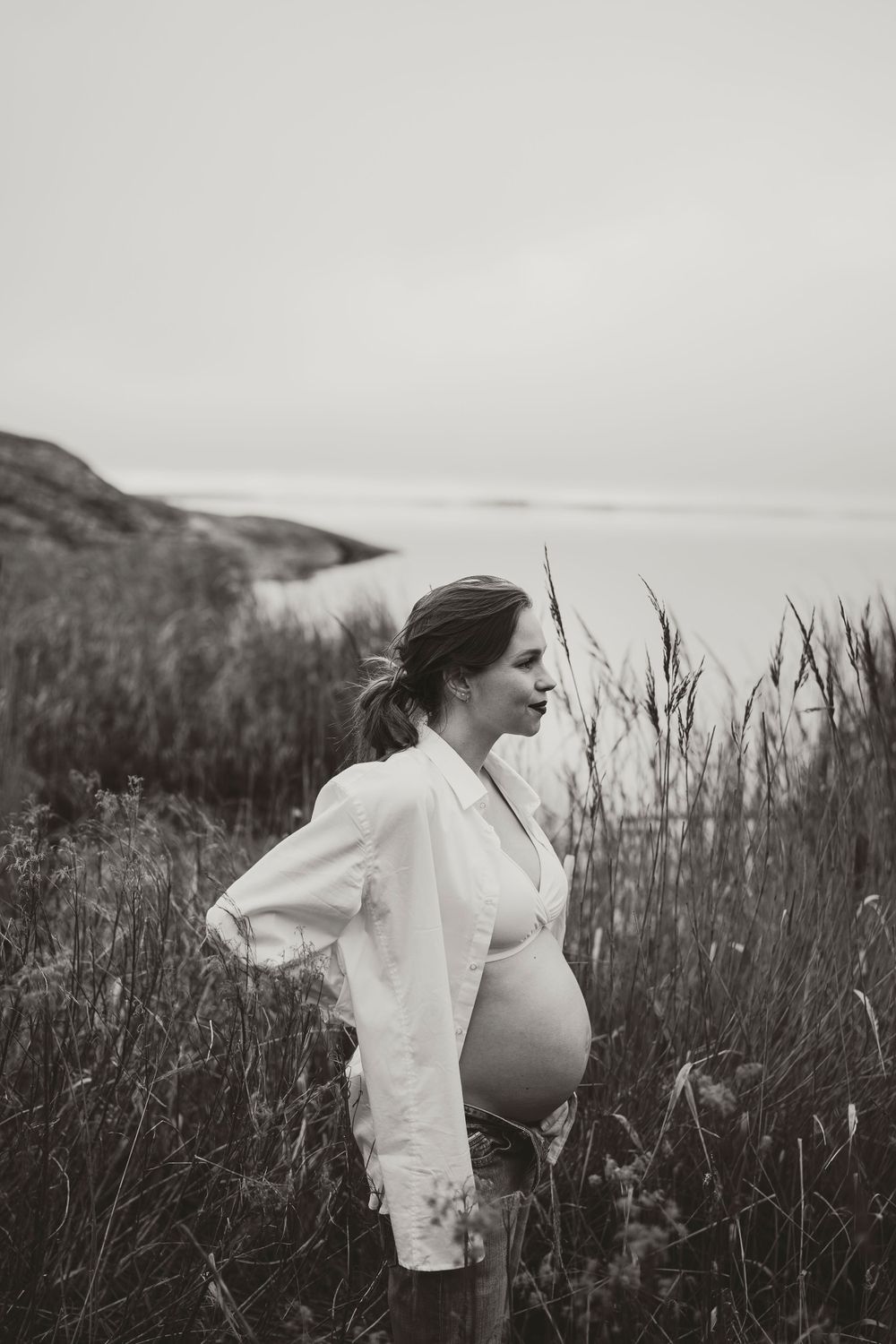 Artistic black and white maternity photo with subject in white shirt against coastal backdrop.