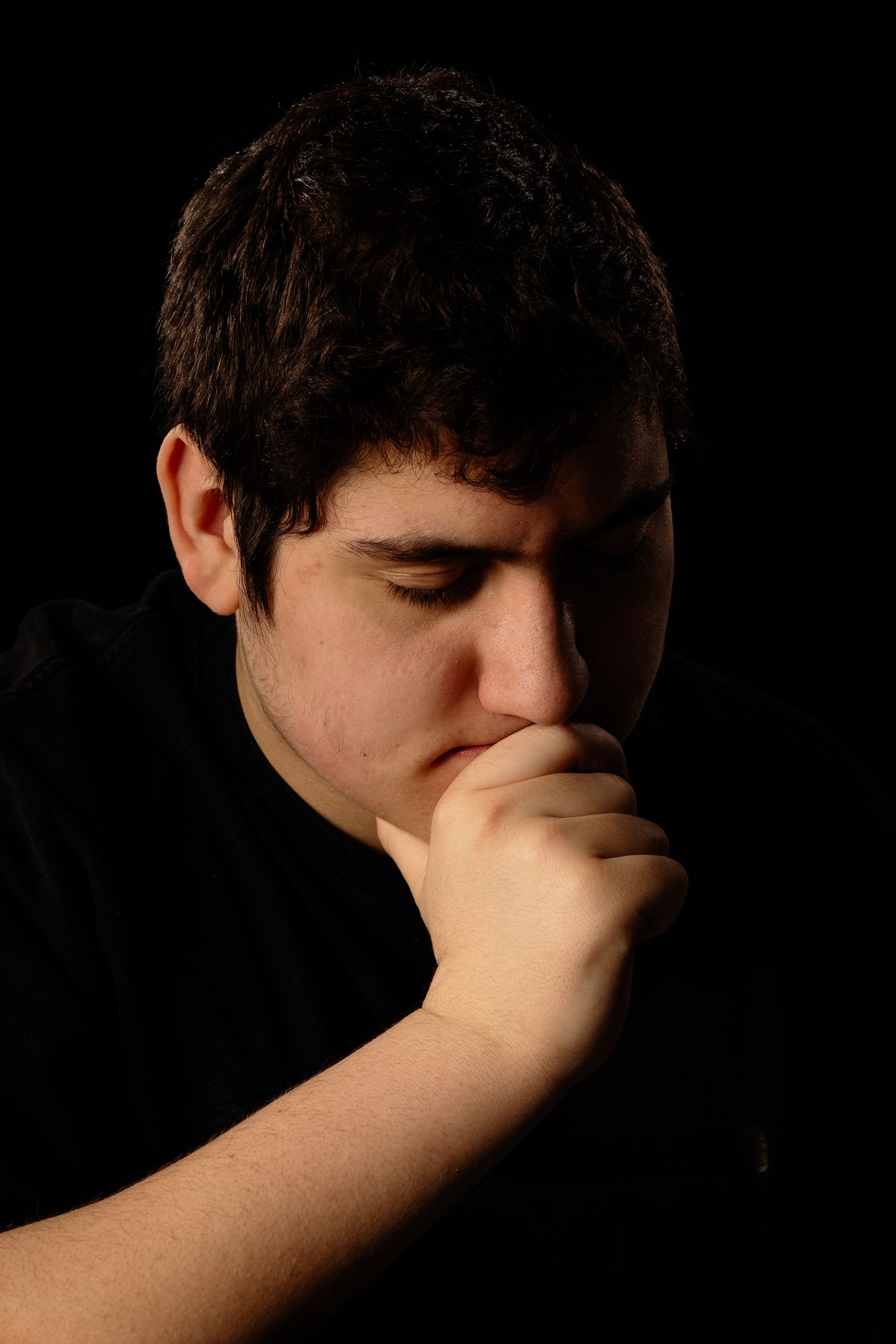 Dark moody portrait of someone in black clothing with hand on chin looking downward.