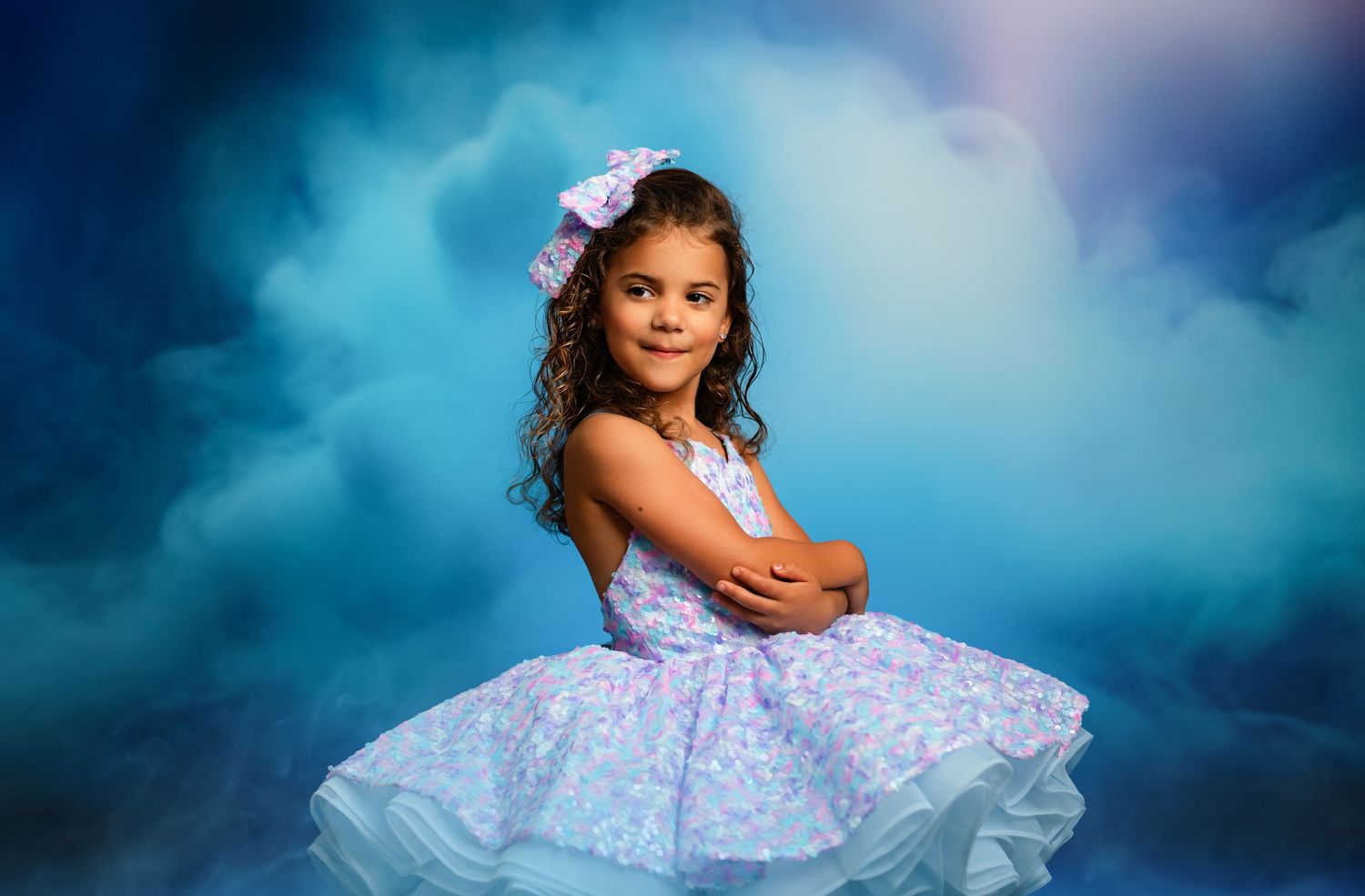 Young model in flowing pink and lavender tulle ball gown poses against purple backdrop in elegant portrait session.