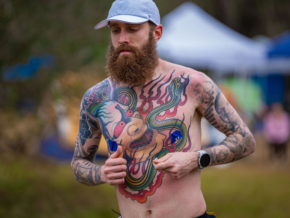Shirtless trail runner with colorful chest tattoos running through aid station area.