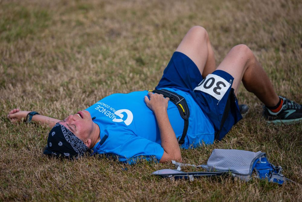 Exhausted runner lies on ground after race wearing blue shirt and race bib.
