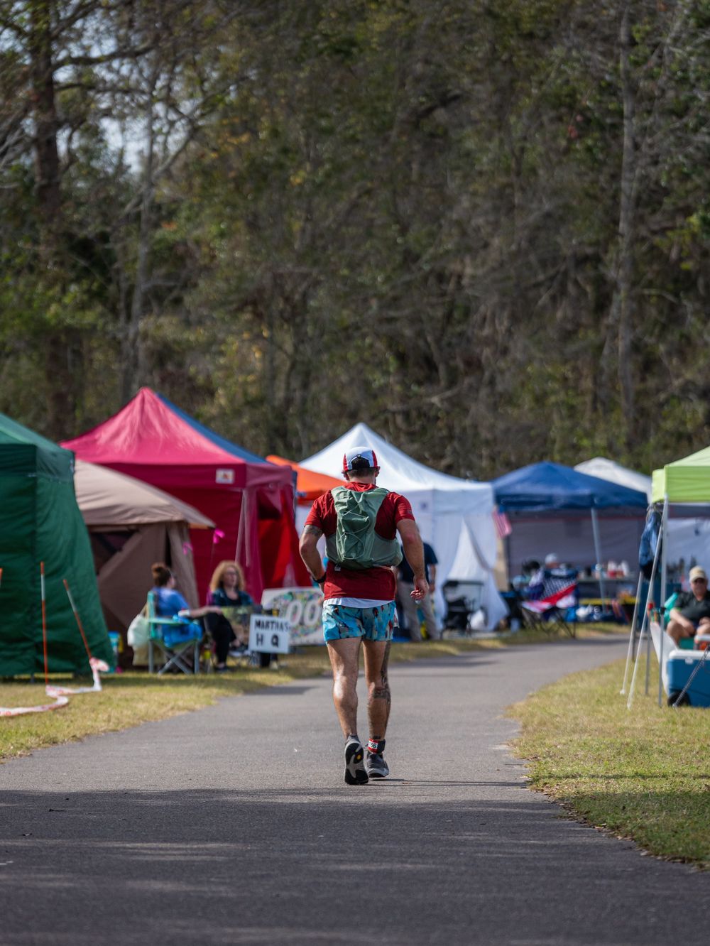 Runner walking past colorful pop-up tents and canopies at an outdoor event on a paved path through wooded area.