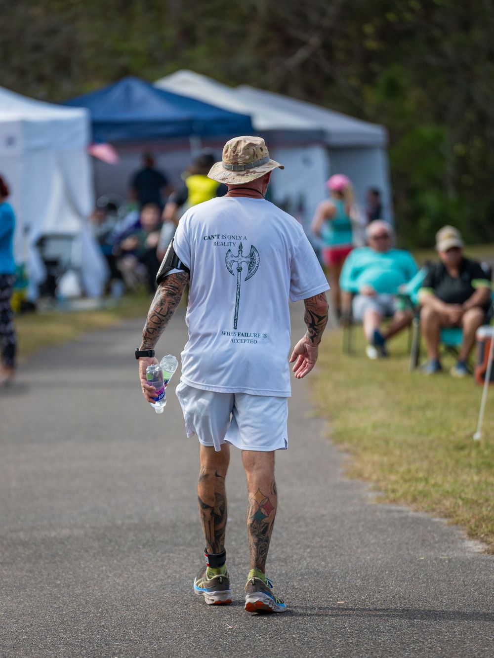 Runner in white shirt and shorts walking along path with tents and spectators in background.