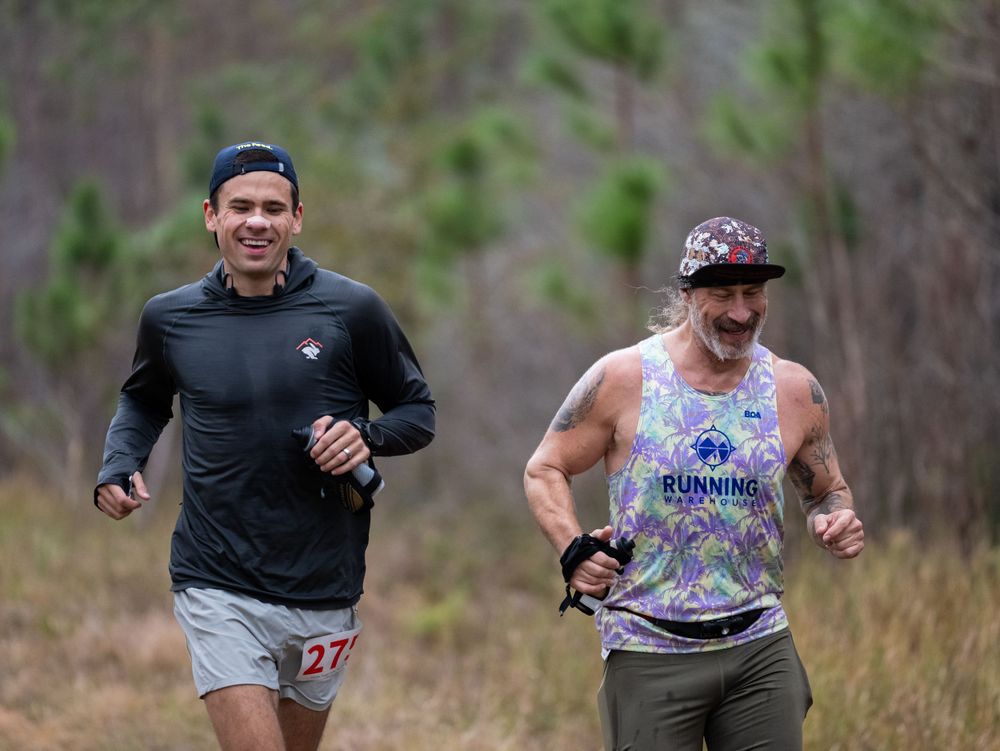Two runners in athletic gear compete on a wooded trail during an endurance race.