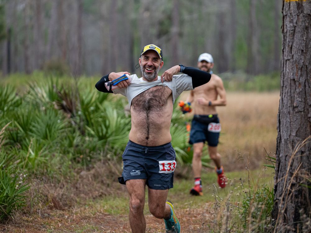 Runner shirtless in trail race through wooded area wearing race bib and holding water bottles.