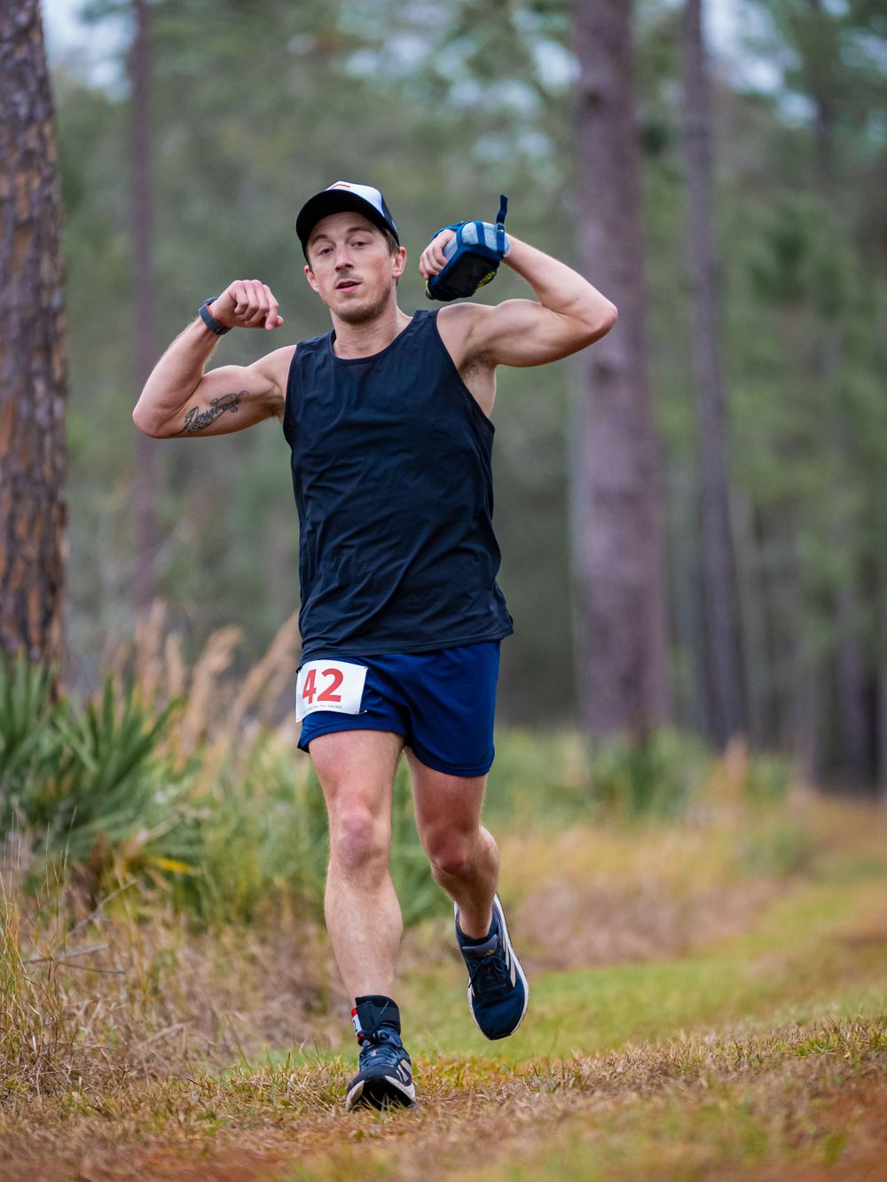 Trail runner in blue tank top and shorts running through forest path with water bottles.
