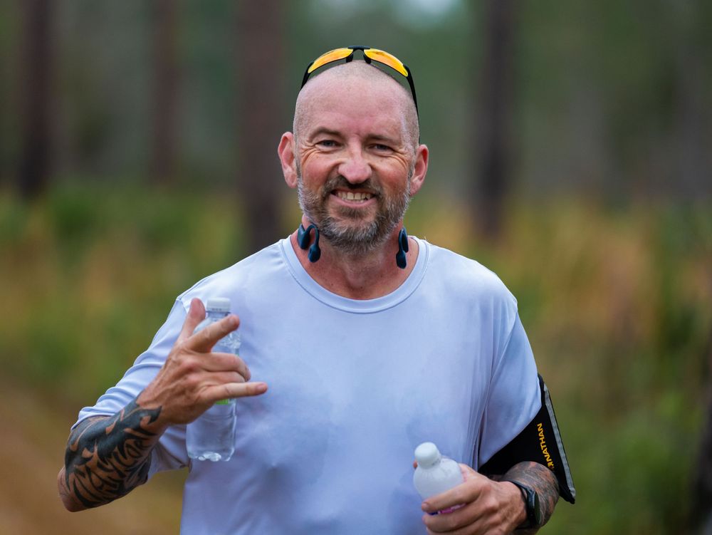 Runner in white shirt and sunglasses gives thumbs up while running through forest trail.