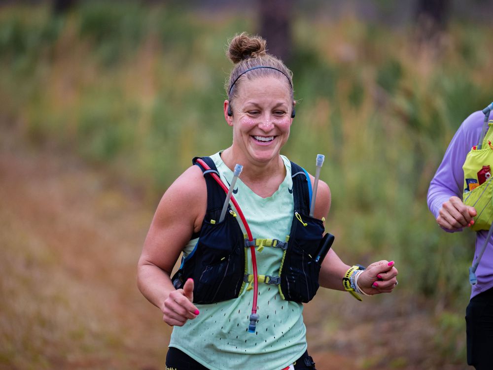 Runner in mint green tank top smiles while competing in trail race wearing hydration vest.