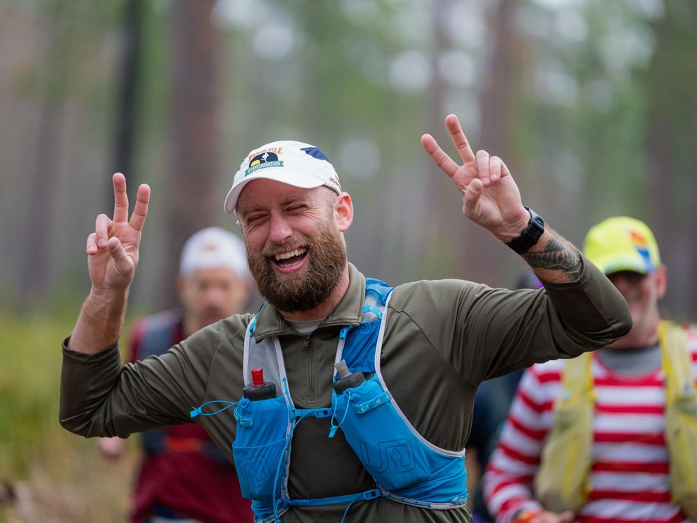 Runner in gray shirt and blue hydration vest gives peace signs while running through wooded course.