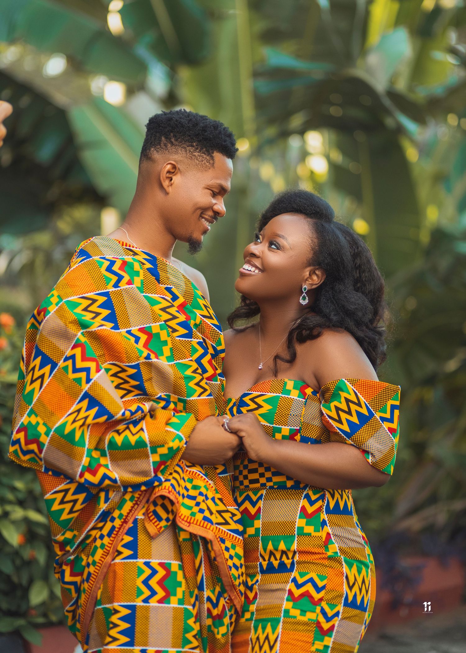 A couple in matching vibrant African kente print outfits embrace and smile against a tropical foliage background.