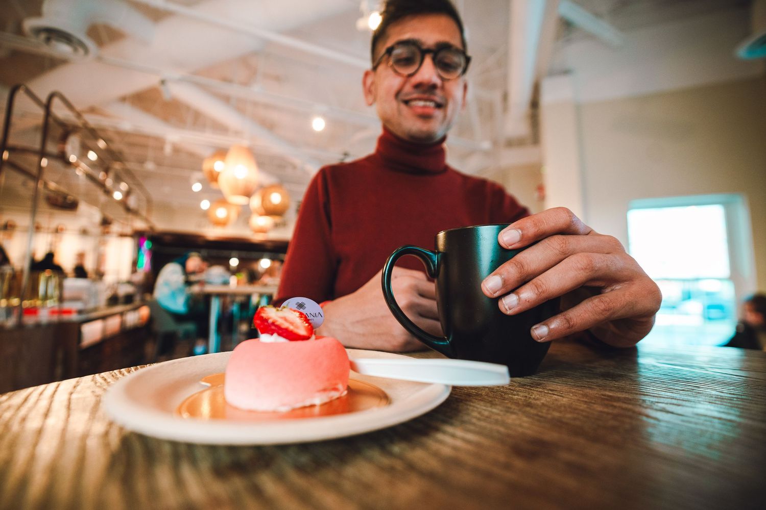 Person in a burgundy turtleneck enjoys coffee and pink dessert at a cozy cafe with warm lighting.
