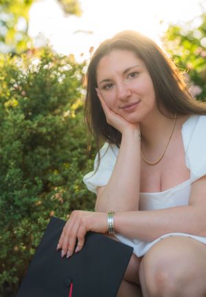 Graduate holds diploma while posing in natural outdoor setting with greenery.