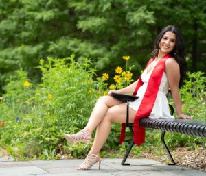 Graduate in red stole poses on park bench surrounded by yellow wildflowers.