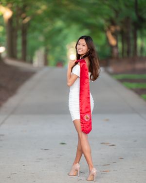 Graduate in a white dress and red stole poses gracefully on a tree-lined pathway.