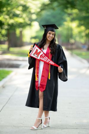 Graduate in cap and gown shows off red graduation stole while posing outdoors.