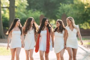 Group of graduates in white outfits and red stoles walking together on campus path.