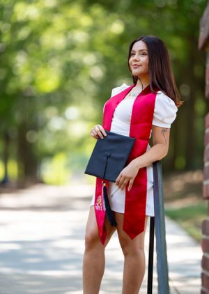 Graduate in red stole poses against railing on tree-lined campus path.