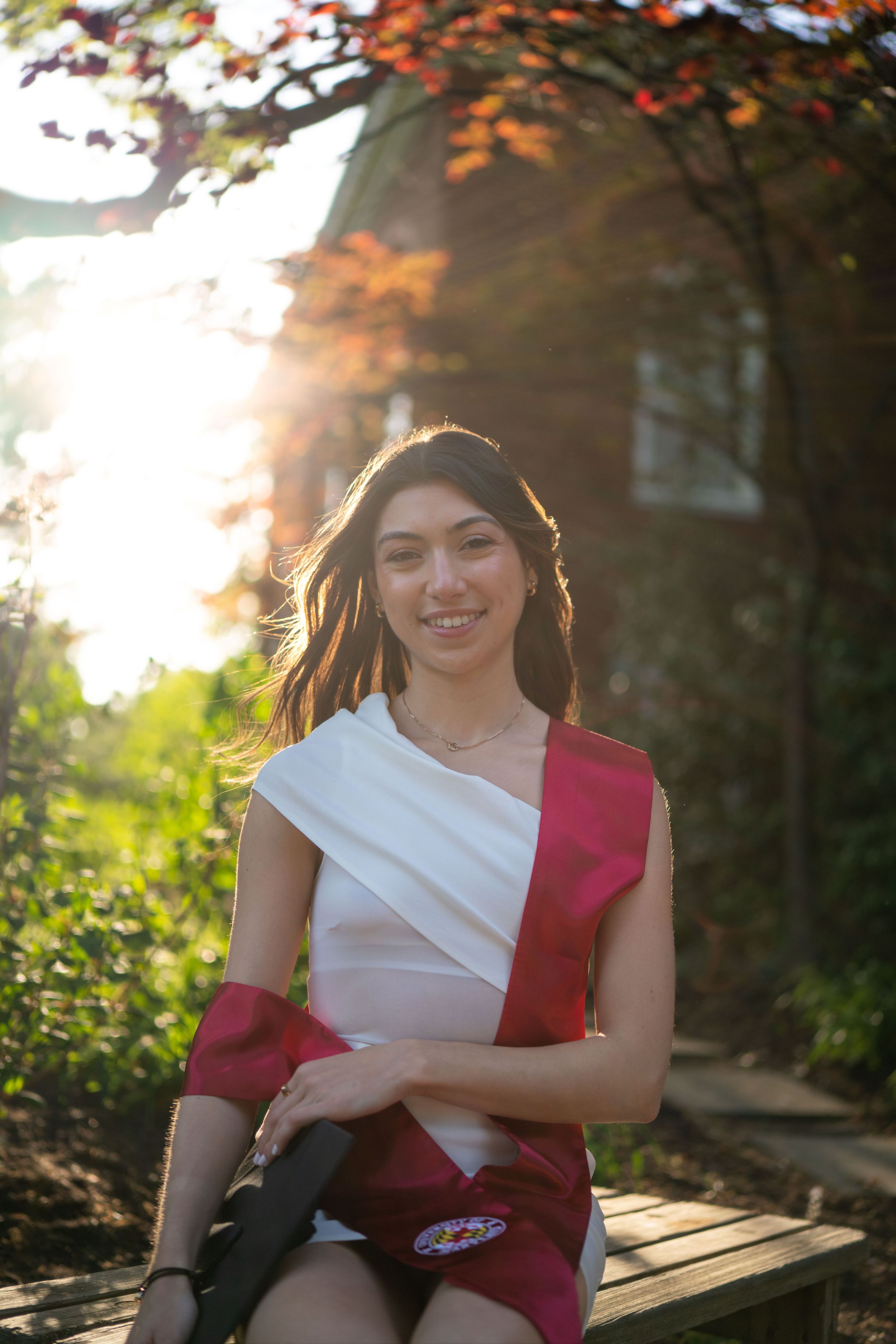 Graduate in red and white stole poses on wooden bench during golden hour sunset photo session.