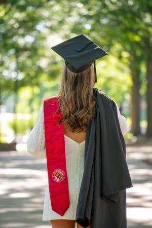 Graduate wearing black cap and red stole photographed from behind on tree-lined path.