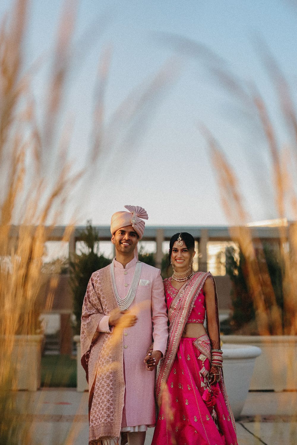 A couple in traditional Indian wedding attire stands together at sunset surrounded by tall grass.