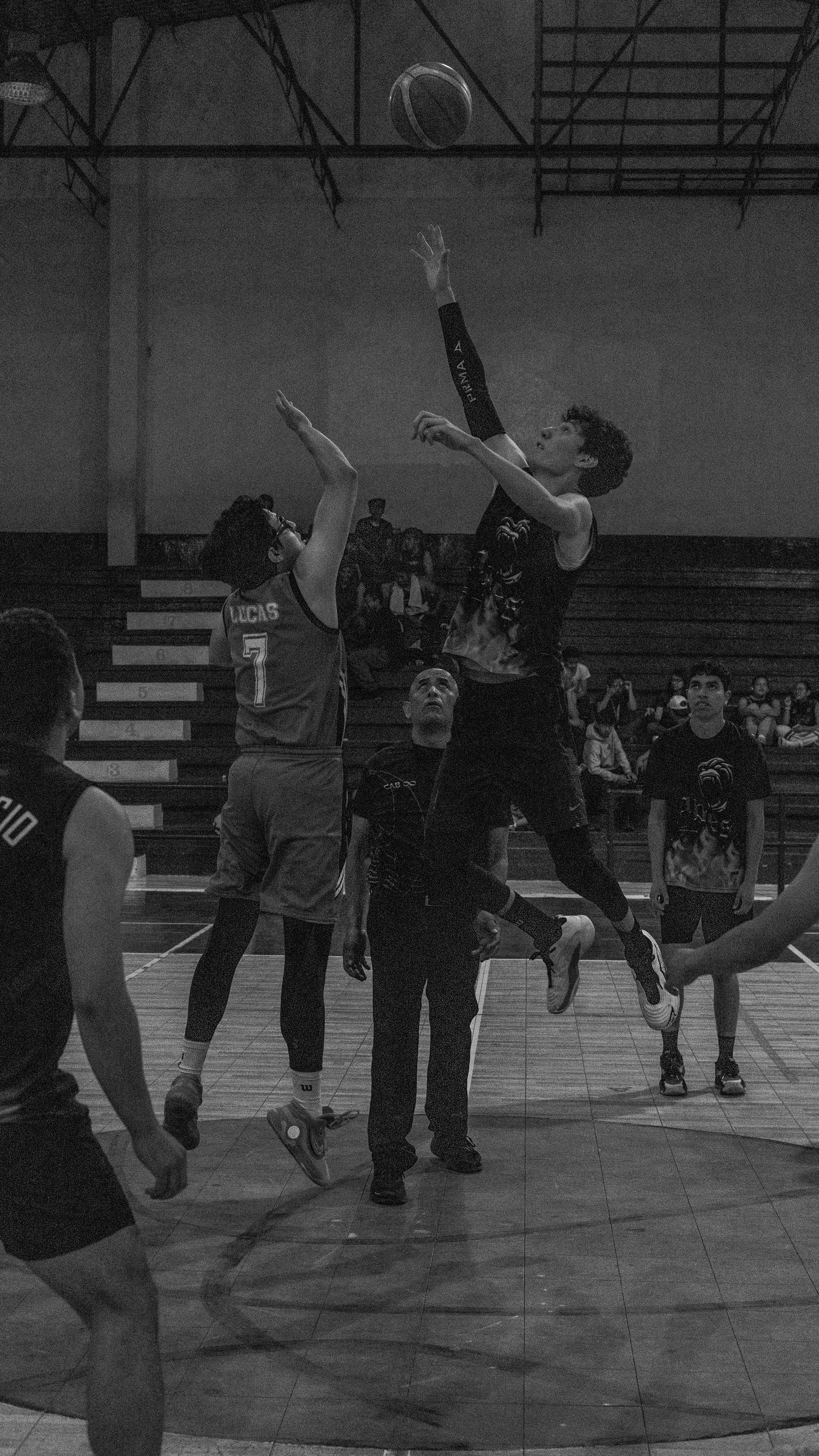 Players jump for the basketball during an intense indoor game captured in black and white.