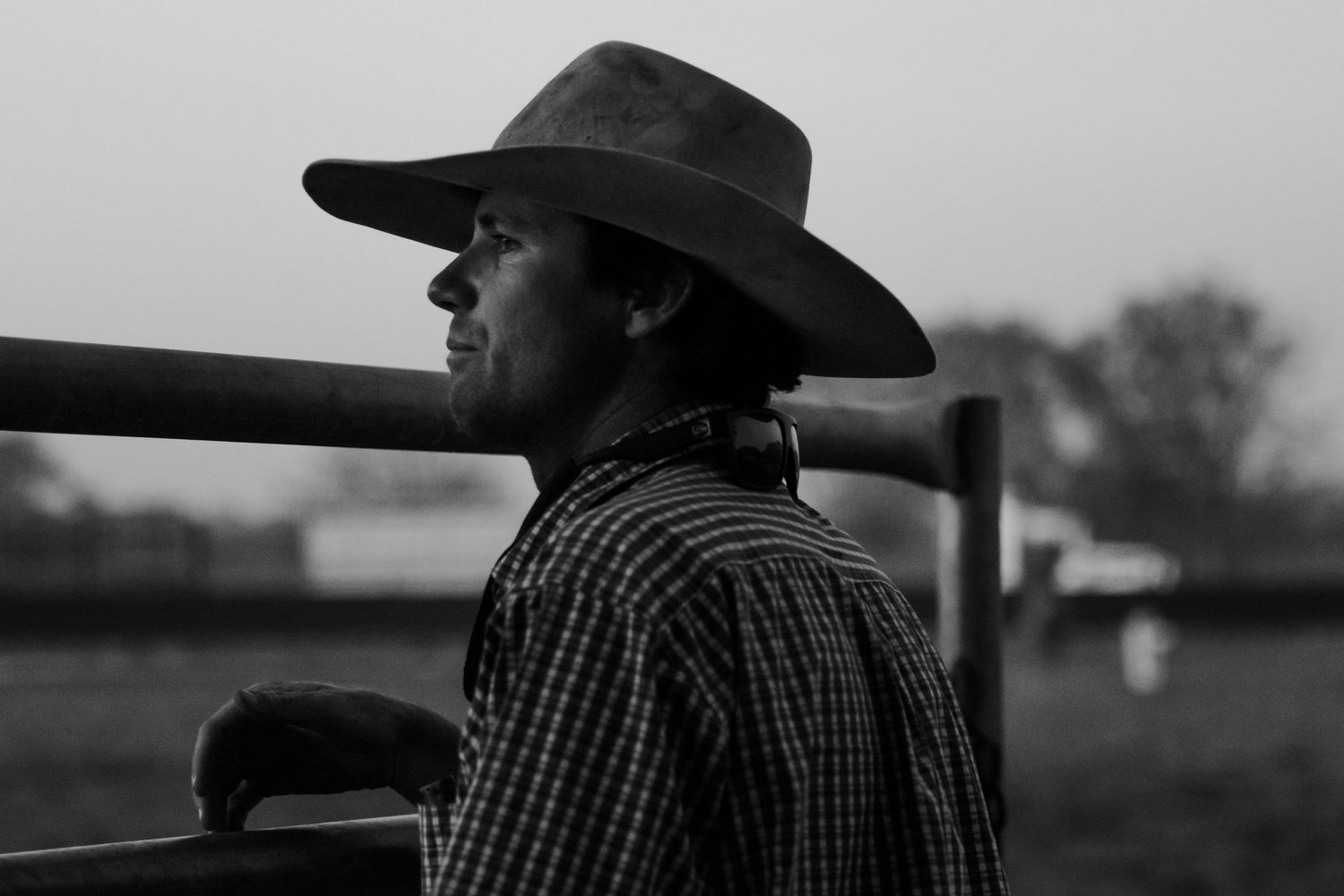 Silhouette of person in cowboy hat leaning against fence rail at dusk.