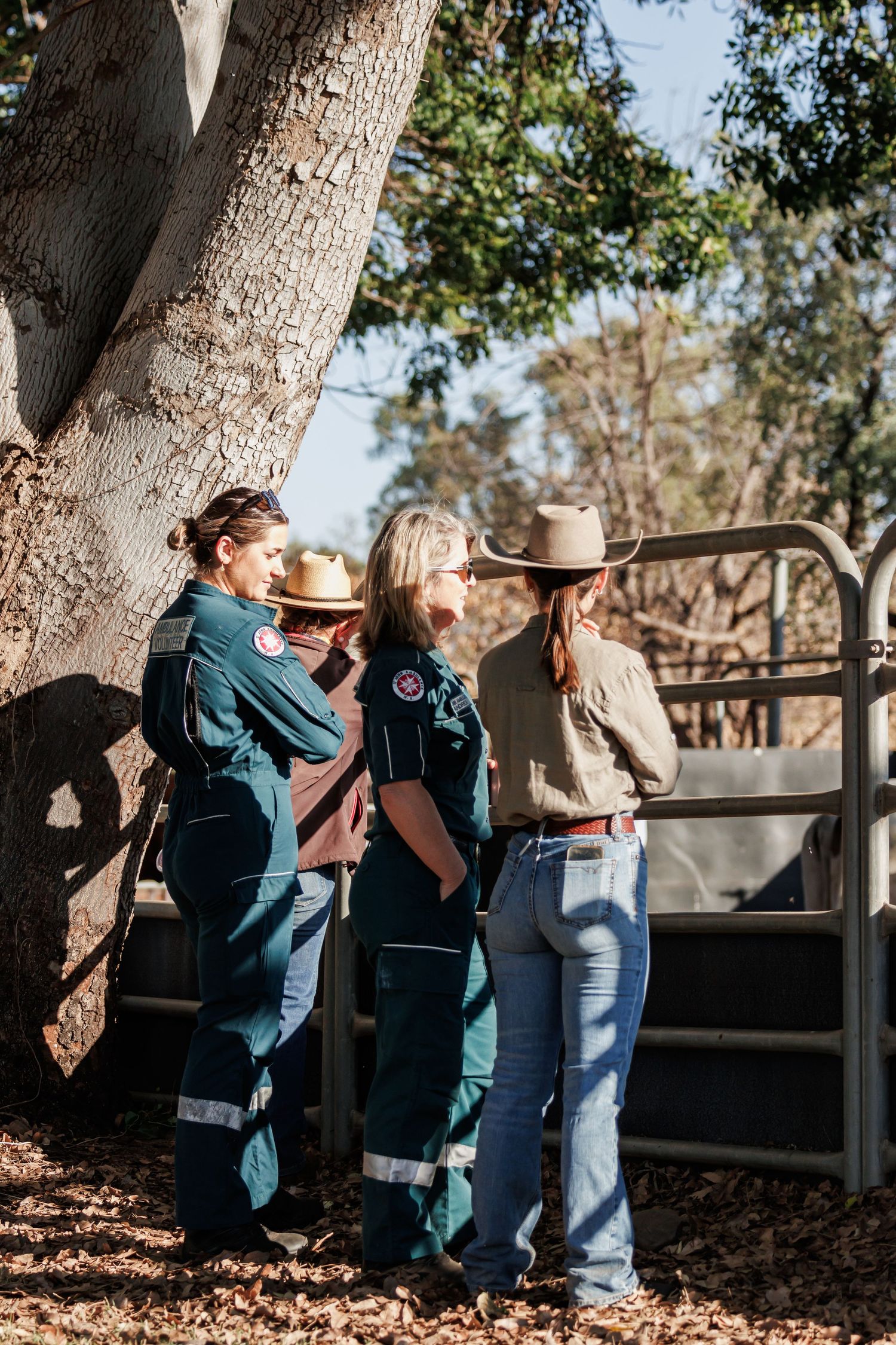 Three people in western attire chat while leaning against a tree near a gate.