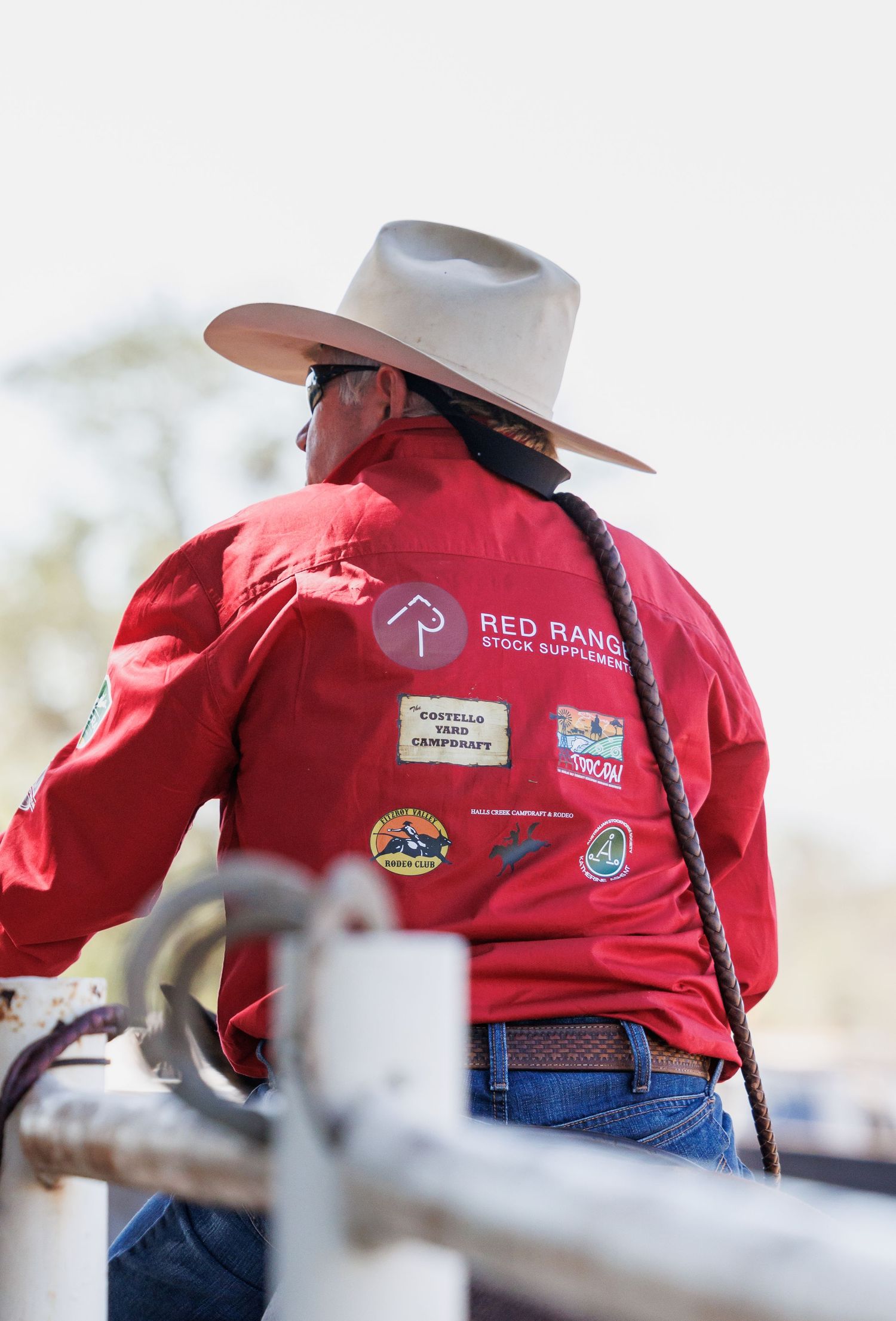 A rider wearing a red shirt with Red Ranch branding stands against a fence.