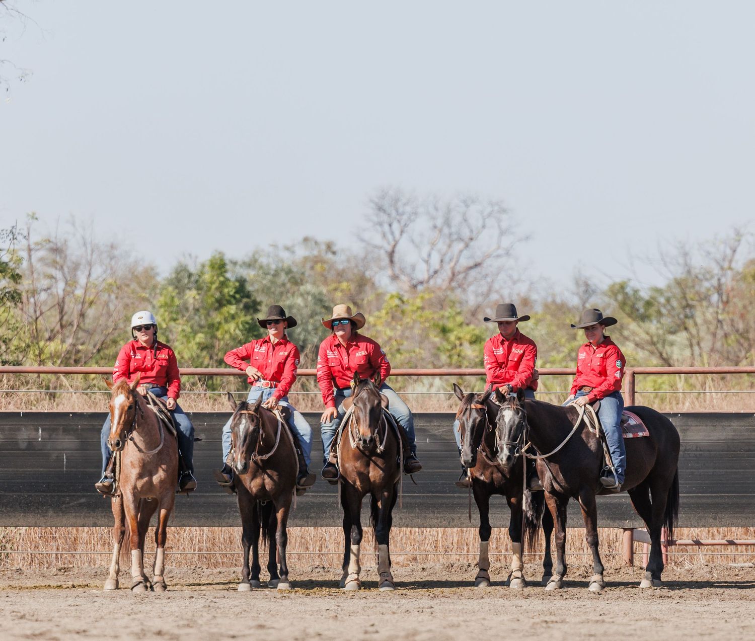Five riders in matching red shirts line up on horseback in formation.