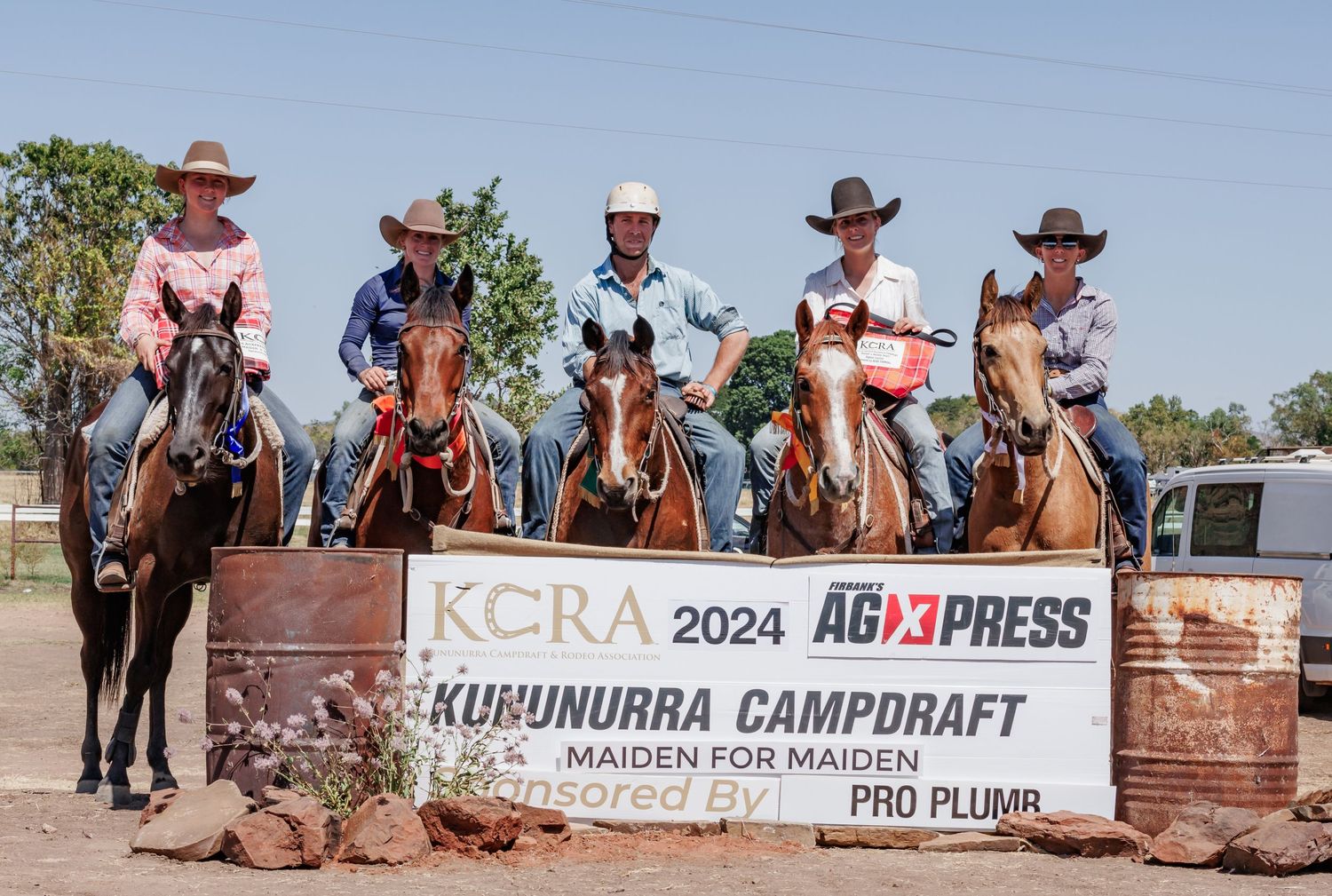 Group of mounted riders pose with competition banner at campdrafting event.
