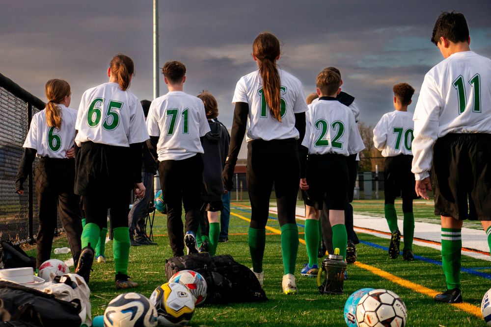 Soccer team lined up on field with numbered jerseys and equipment bags at their feet.