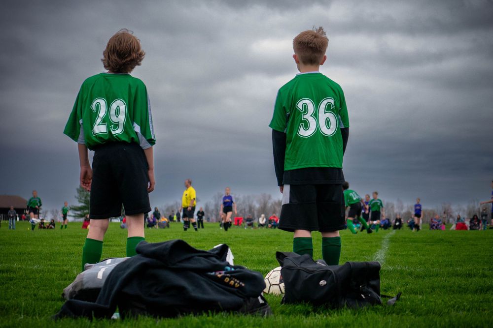 Two soccer players stand near equipment bags under stormy skies.