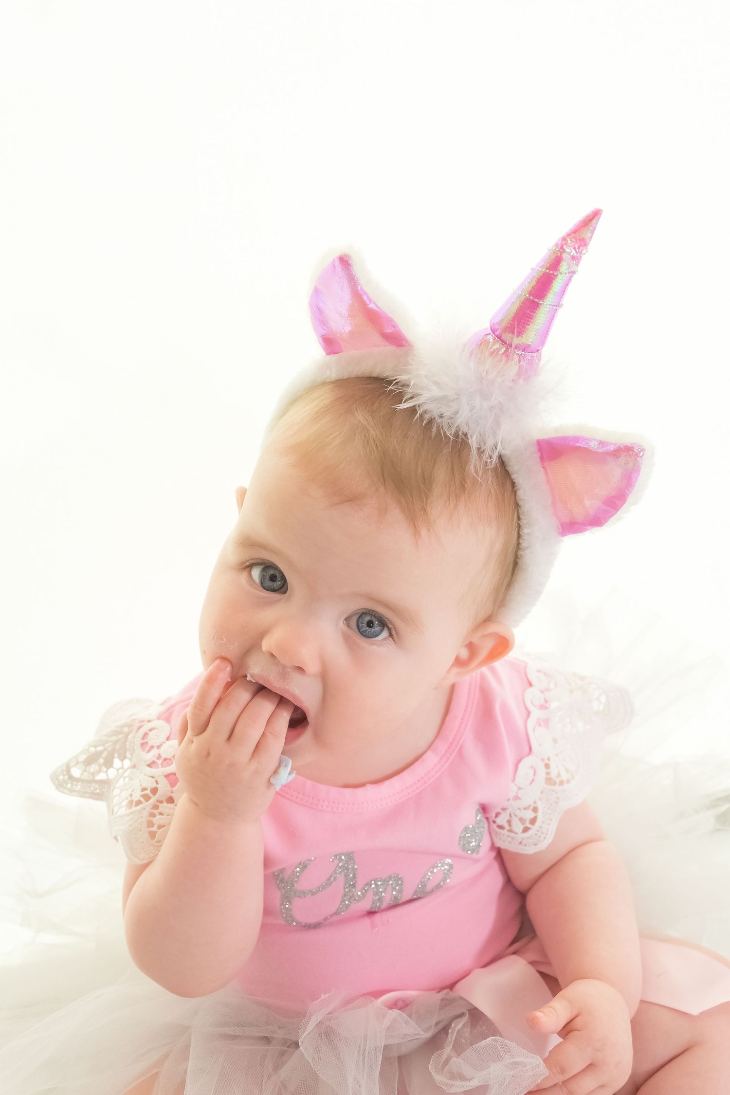 Baby in pink outfit and unicorn headband with hand near mouth.