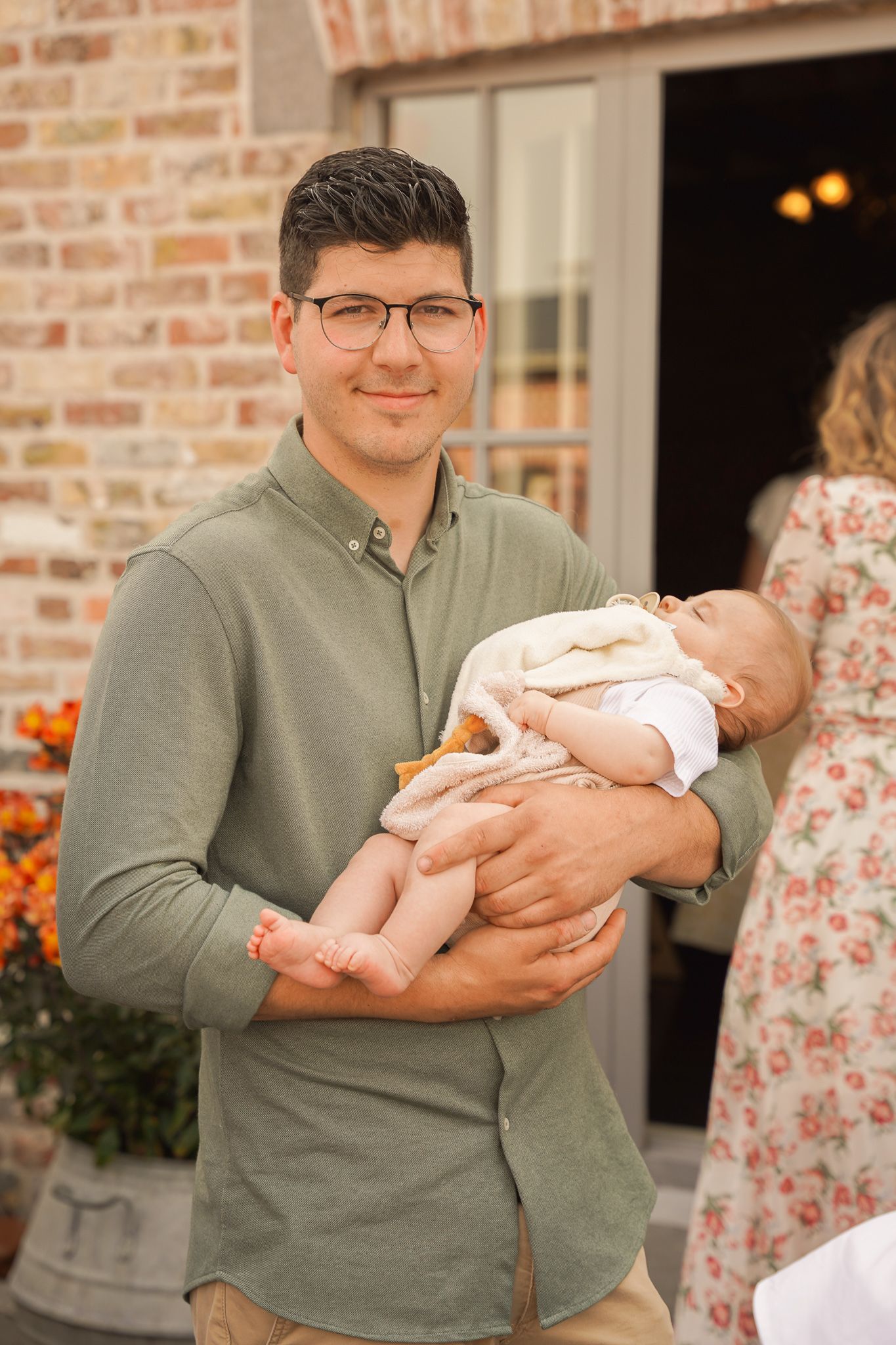 Caregiver holding sleeping infant outside brick house with fall decorations.