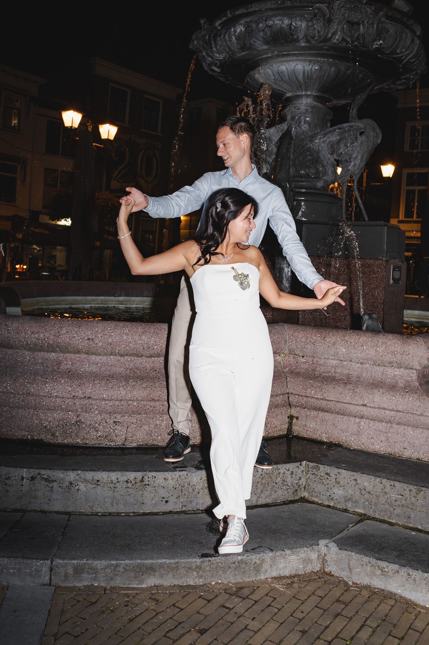 A couple dances playfully at night near an ornate fountain with glowing street lamps in the background.