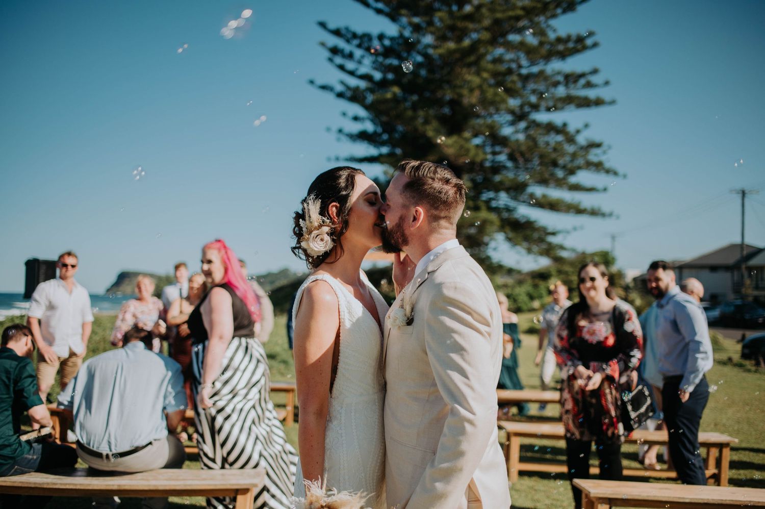 A wedding couple shares an intimate moment during an outdoor ceremony with guests and a pine tree in the background.
