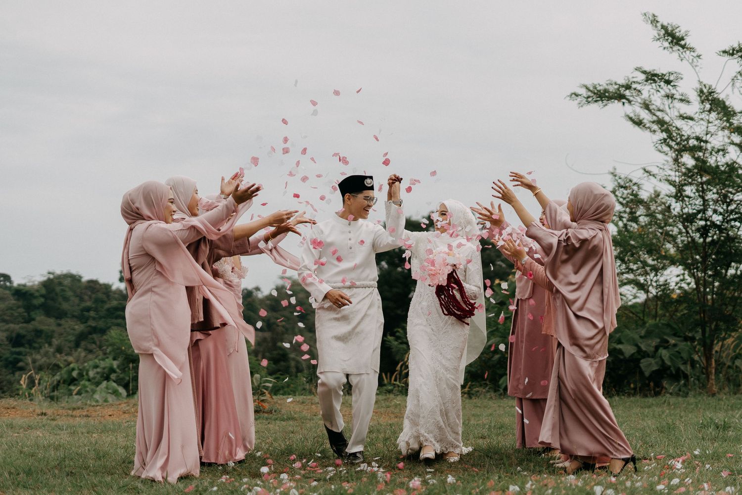 Joyful Malay wedding moment as the bride and groom walk through a shower of flower petals, surrounded by bridesmaids. Hizashi Studio