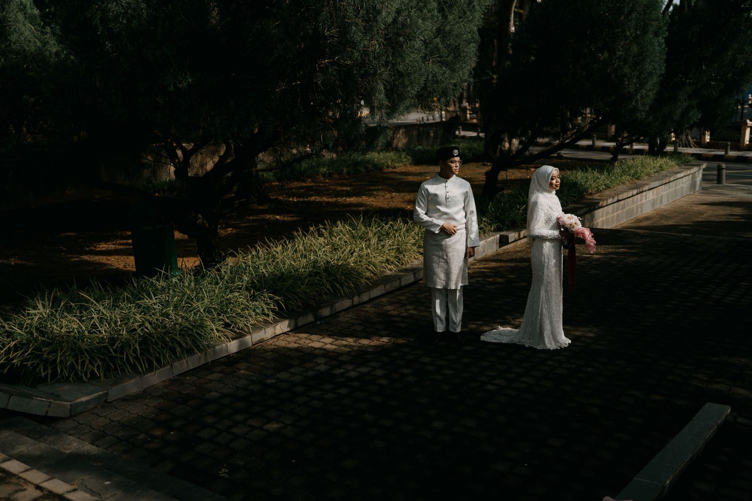 Malay bride and groom in elegant white attire stand in a serene outdoor setting. Captured by Hizashi Studio, Kuala Lumpur & Pahang