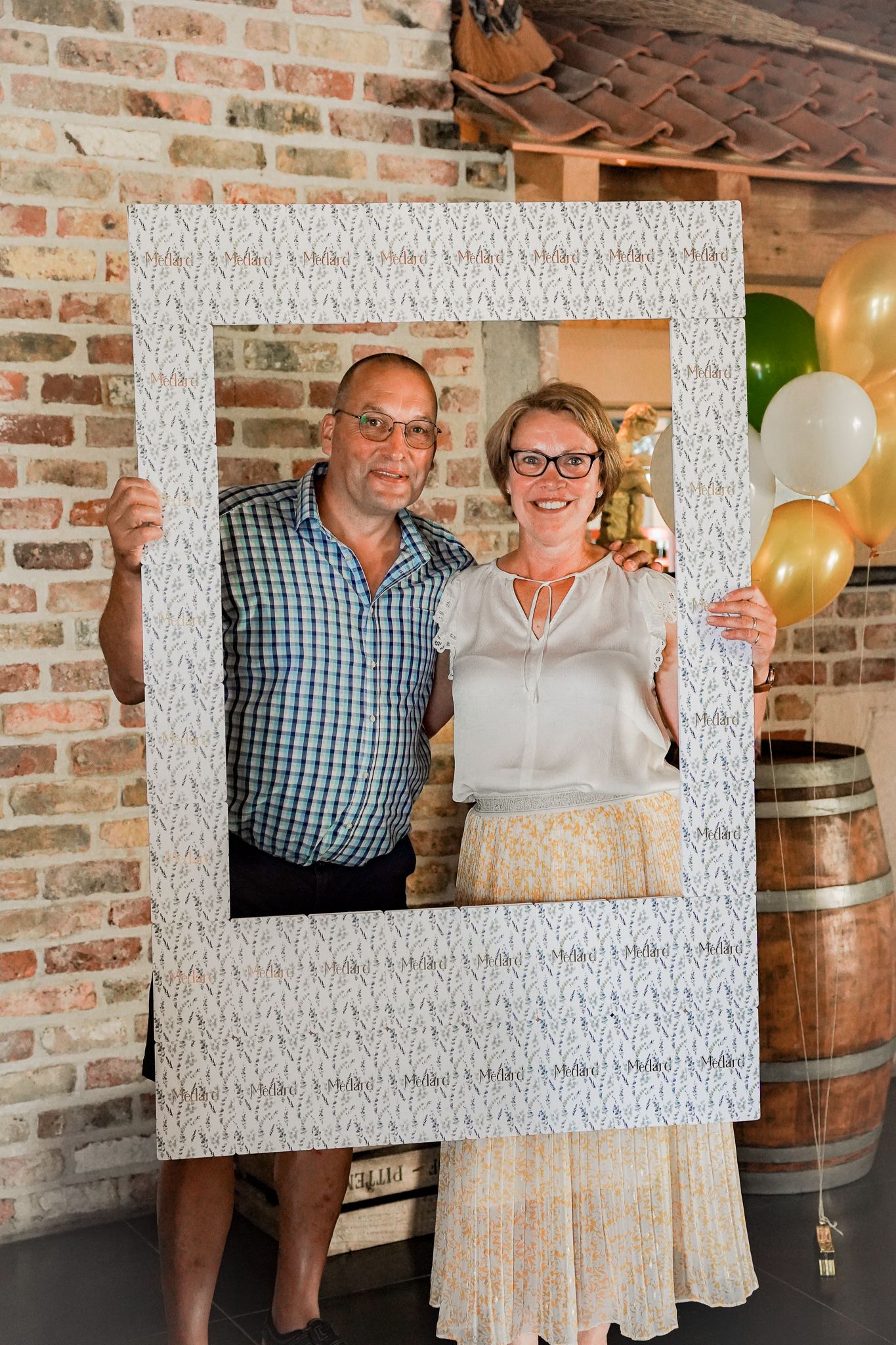Couple posing with oversized white photo frame at party with brick wall and balloons.