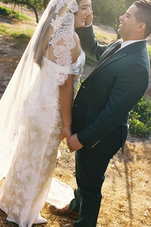 A couple in wedding attire stands close together outdoors, with sunlit scenery behind them.