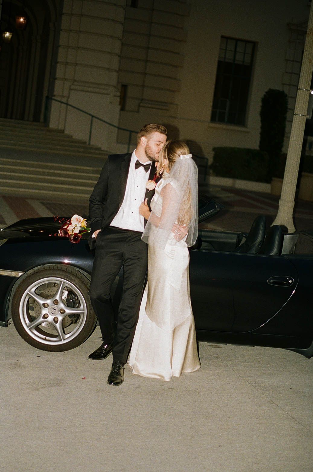 A bride and groom share romantic moments in an outdoor parking lot as a vintage car sits in the background.