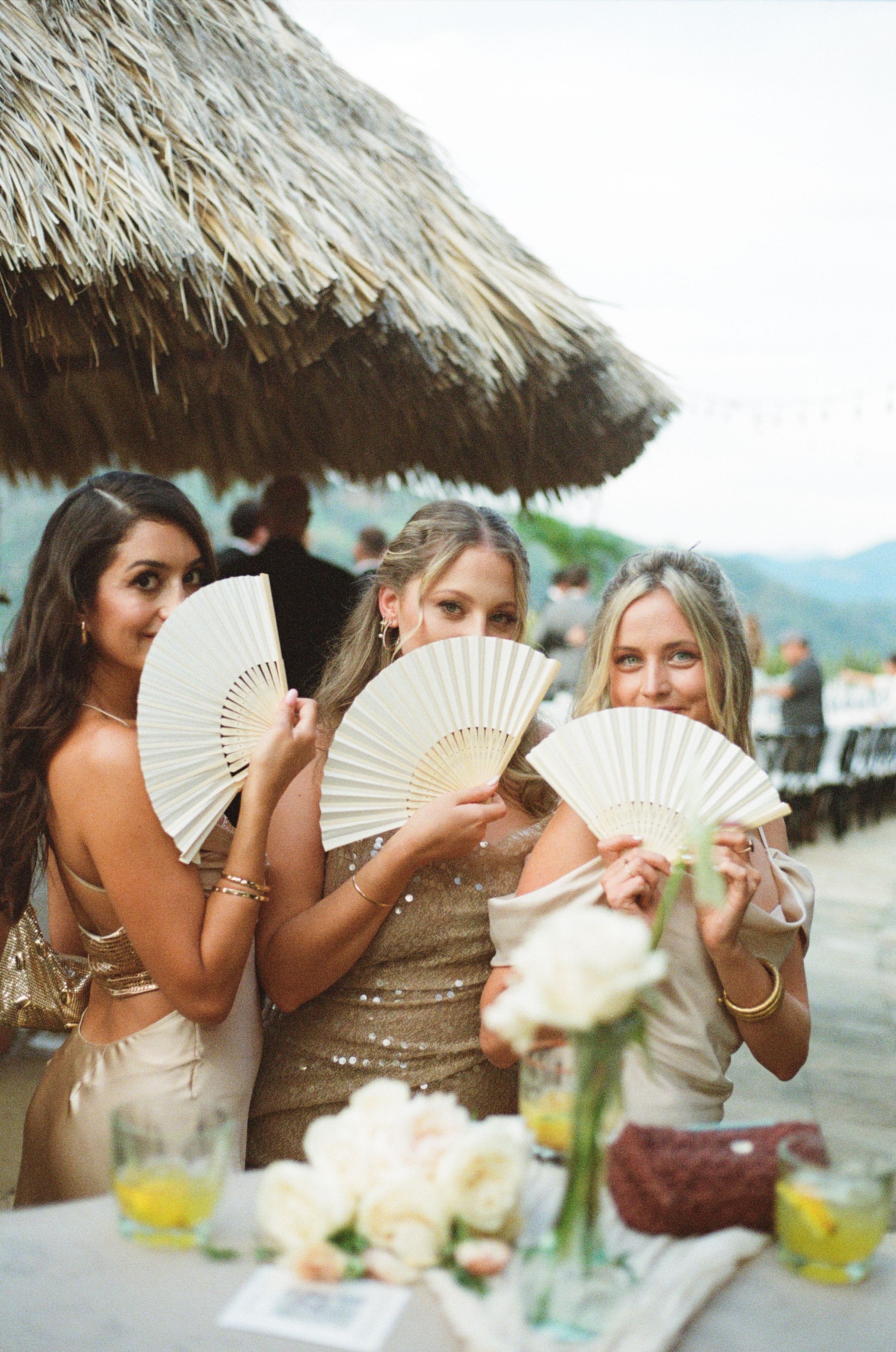 Three attendants in sparkly dresses hold decorative white fans under a thatched roof at a tropical venue.