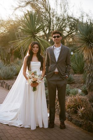 Bride and groom pose together in desert garden with yucca plants and stone pathway at sunset.
