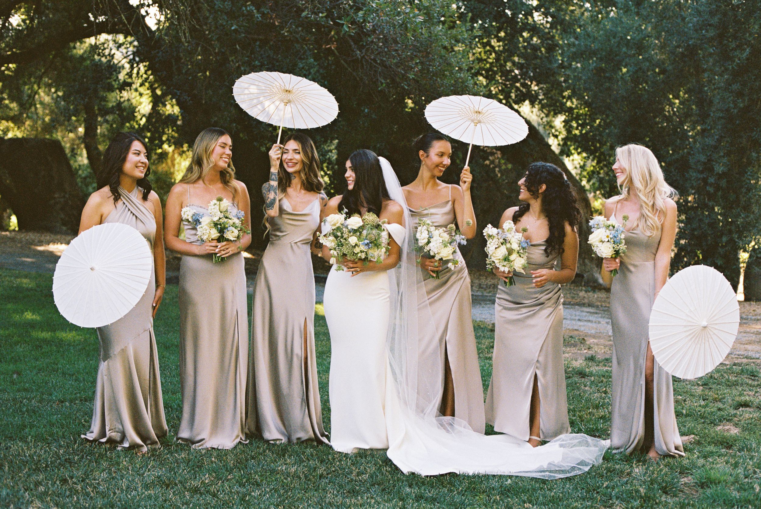 Bridal party in champagne dresses poses with white parasols in garden setting.