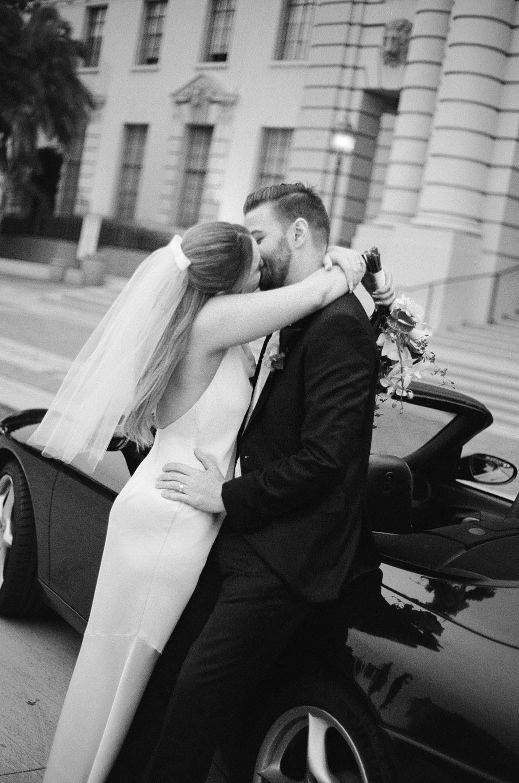 A romantic moment between newlyweds next to a luxury car outside a classic building.